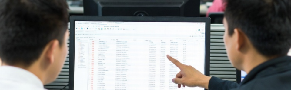 Two business people looking at a screen, one pointing to data sheet